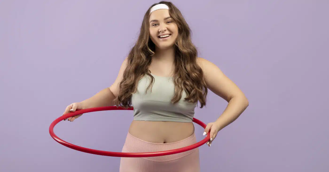 Smiling young European plus size female in uniform doing exercises with hula hoop